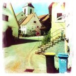 Neben zwei Mülltonnen führt eine gebogene Straße aufwärts zwischen Dorfhäusern hindurch auf eine kleine Kirche hinzu.