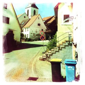 Neben zwei Mülltonnen führt eine gebogene Straße aufwärts zwischen Dorfhäusern hindurch auf eine kleine Kirche hinzu.