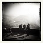 Schwarz-weiß-Aufnahme eines Aussichtspunkts über drei Personen, die vor einer kleinen Mauer stehen und über diese hinweg ins dunstige, gegenlichtige Tal blicken.