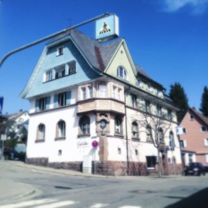 An langem Galgen hängt ein Zebrastreifenschild mitten über einer kleinstädtischen Kreuzung. Dahinter ein schwarzwäldisches Stadthaus mit Krüppelwalm und großem Erker.