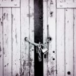 Schwarz-weiß Foto eines Brettertors, das mit einer Kette und Vorhängeschloss verschlossen ist. Detailaufnahme.