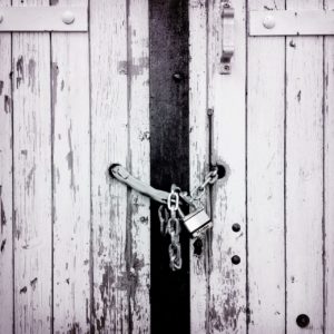 Schwarz-weiß Foto eines Brettertors, das mit einer Kette und Vorhängeschloss verschlossen ist. Detailaufnahme.