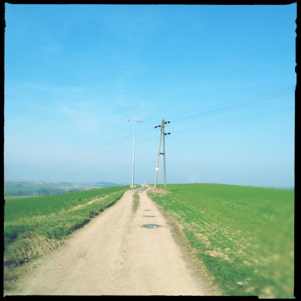 Ein Feldweg führt durch hellgrüne Felder schnurgerade auf einen Stommast zu. Darüber blauer Himmel.