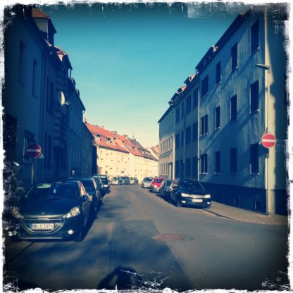 Die gewundene Stadtstraße führt durch lückenlos bebaute dreistöckige Wohnhäuser. Im schrägen Sonnenlich schimmert die rechte Häuserphalanx.