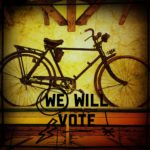 Gelblich schimmernder Hintergrund mit einem alten Fahrrad im Profil. Darunter der Schriftzug We Will Vote