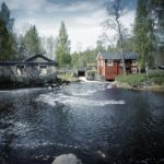 Ein Bach mündet über ein kleines Wehr zwischen einem roten Holzhaus und einem Steinernen Bau in einen aufgewühlten, schwarzblauen See.
