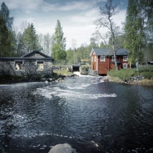 Ein Bach mündet über ein kleines Wehr zwischen einem roten Holzhaus und einem Steinernen Bau in einen aufgewühlten, schwarzblauen See.