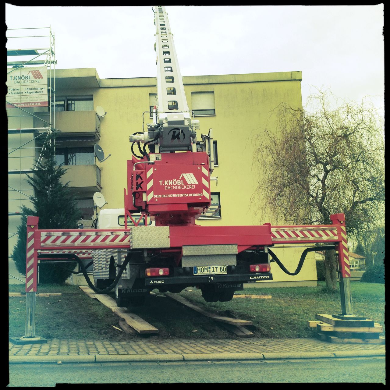 Ein fahrbarer Krand mit ausgefahrenen hydraulischen Stützen vor einem Flachdach-Mietshaus. Das rote Fahrzeug scheint wie zu fliegen. Die Räder hängen in der Luft.