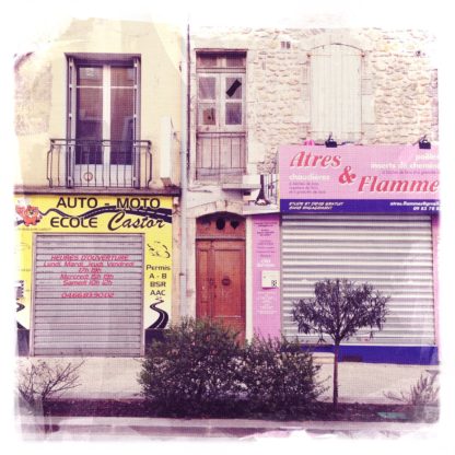 Fehlfarben Retrobild einer kleinstädtischen Häuserfront in Frankreich. Die Fenster der Läden sind mit schweren, eisernen Rolläden verhangen. Man liest Auto-Moto Ecole Castor und Atres & Flamme