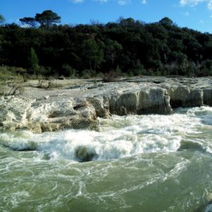 Die beiden unteren Drittel des Bildes zeigen Kaskaden und Felsen in Grau und weiß mit wildem Wasser. Im Hintergrund teilen sich dunkelgrüne Pinien und blauer Himmel das obere Drittel.