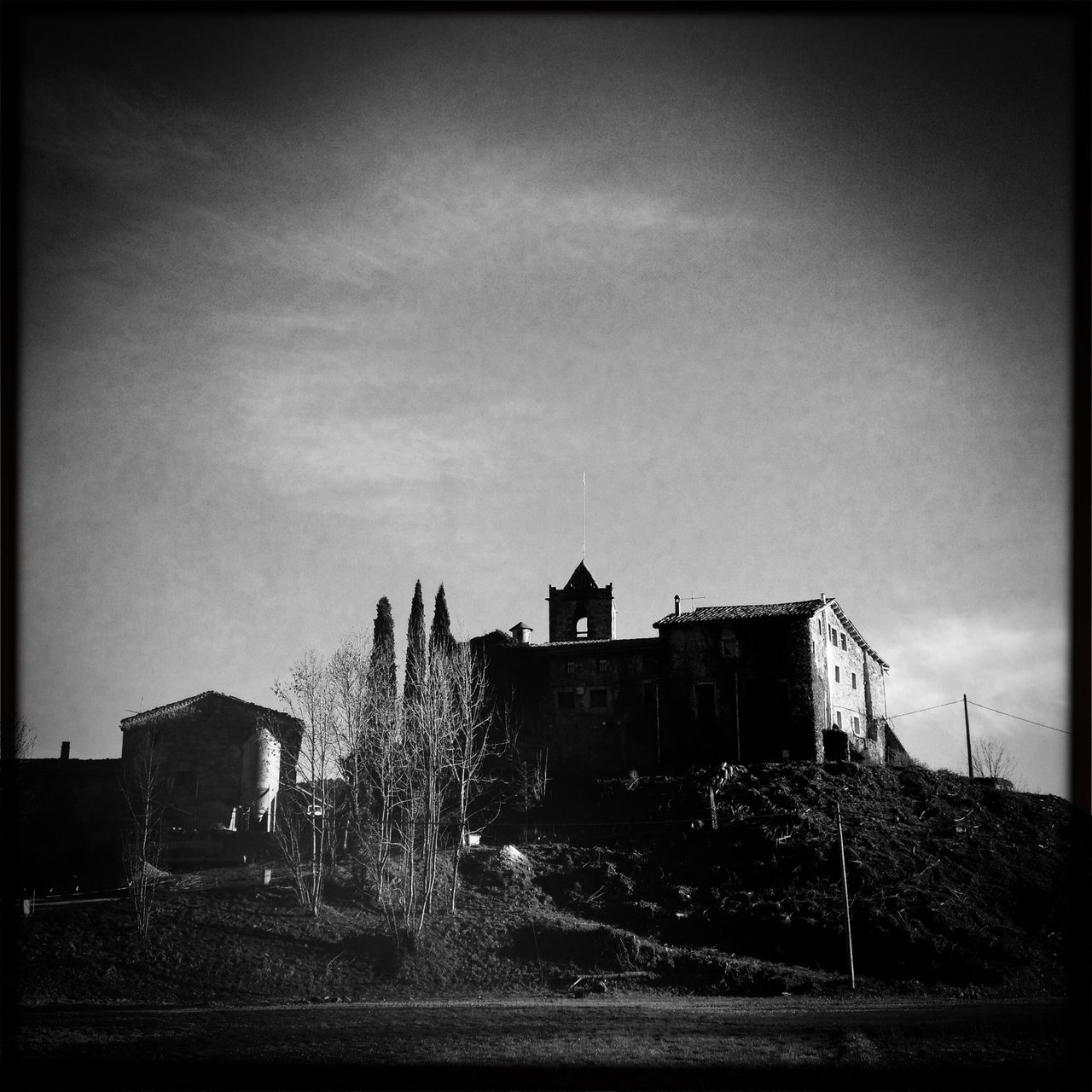 Schwarz-Weiß-Bild einer katalanischen Kirche ein bisschen Underfoot aufgenommen im Gegenlicht. Dunkle Vignette an den oberen Bildecken. Kahle Bäume geleiten den Blick.