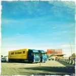 Autobahnrastplatz in Siebziger hellblau-gelb-Fehlfarben. Zwei LKW nebeneinander vor einem Hotel. Betonblockaden führen den Betrachter zum Hoteleingang. Der blaue Himmel nimmt fast drei Viertel der Bildfläche ein.