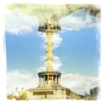 Die Berliner Siegssäule gespieglt und in ihren eigenen Kopf kopiert, so dass ein Streifen Himmel mit Schönwetterwolken zwischen den oben und unten liegenden Säulengebäudem am Sockel der Säule liegt wie die Spiegelung in einem See.