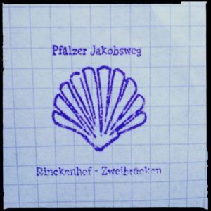 Blauer Stempel auf kariertem Papier, etwas unscharf. Aufschrift Pfälzer Jakobsweg über einer skizzierten Jakobsmuschel und darunter die Schrift Rinckenhof-Zweibrücken.