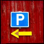 Parkplatzschild über gelbem, nach links zeigendem Pfeil an roter Holzbretterwand.