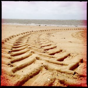 Riesige, Fischrätenartige Traktorreifenspur im Sand vor grauem Meer.