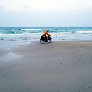 Schwer bepackter Reiseradler mit gelber Regejacke vor verregnetem Meer auf Sandstrand radelt von links nach rechts.