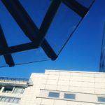 Wie ein nlandendes UFO schiebt sich die gläsern-stählerne Konstruktion des Dachs eines Pavillons in den blauen Himmel. Im hintergrund die leicht überbelichtete Fassade eines mehrstöckigen Krankenhausgebäudes.