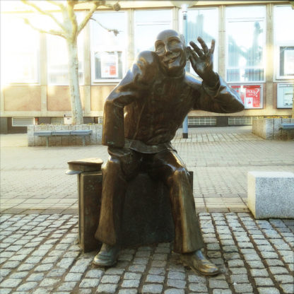 Eine sitzende Bronzefigur, lebensgroß auf innerstädtischem Platz, hält die linke Hand lauschend ans Ohr und reckt markant den Kopf.