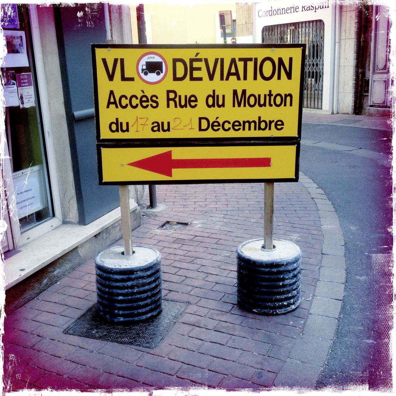 Französische Sprache auf einem großen, gelben, rechteckigen Umleitungsschild auf einem innerstädtischen Gehweg, der durch das Schild fast vollständige verstellt wird.