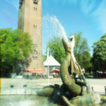 Eine sich aufbäumende Drachenfigur in einem Brunnen speiht Wasser. Im Hintergrund des sommerlichen städtischen Platzes steht ein quadratischer Backsteinturm.