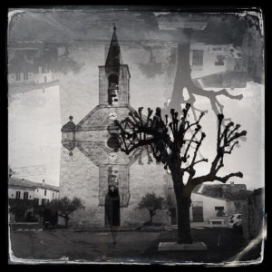 Schwarz-weiß-Bild einer französischen Dorfkirche mit verstümmelter alter Platane als Spiegelung, die wie eine Fatamorgana wirkt. Die Turmuhr zeigt 15:44 Uhr.
