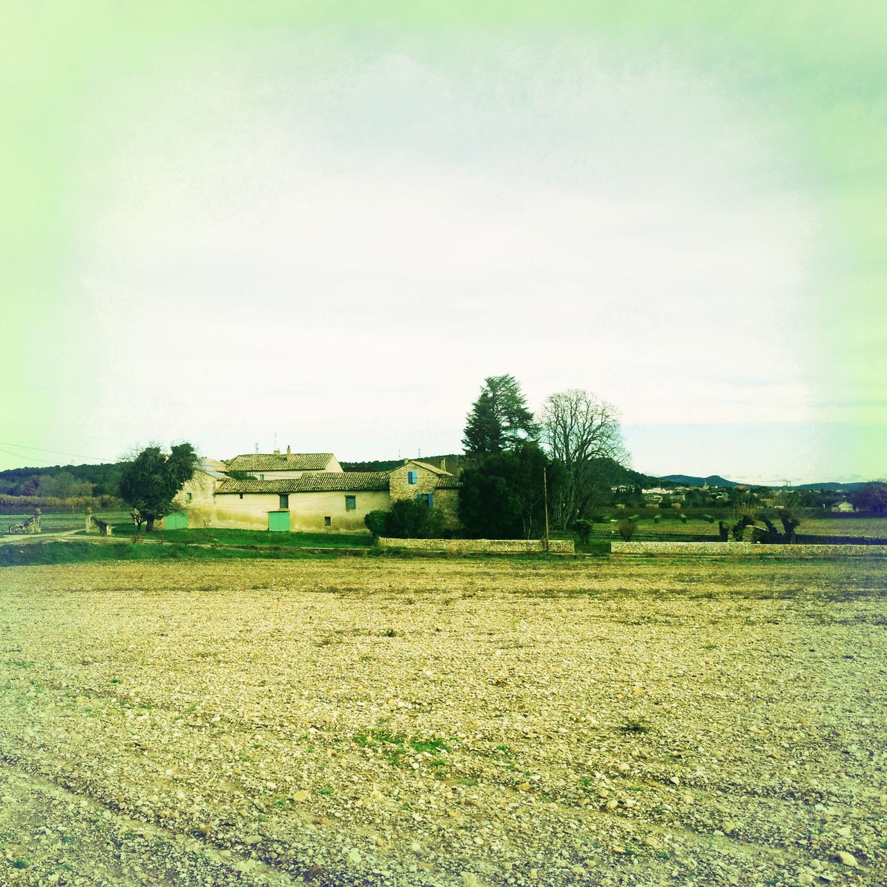 Ein provencalisches Landgut, grün-gelblich fehlfarben unter cyanfarbenem Himmel. Kahles Feld davor.