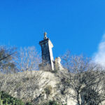 Einen felsigen, von winterlich südfranzösischem Bewuchs gezierten Hang hinauf blicktman auf einen quadratischen Turm, auf dessen Flachsach eine Figur thront. Viel blauer Himmel.