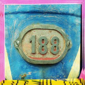 Bunter Rahmen gelb und rosa um eine Zahl 188, die als Alurelieffplatte an einem blauen Hintergrund verschraubt ist.