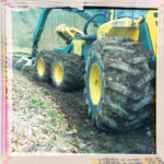 Gelblich-bläuliche Forstmaschine mit drei Rädern und Hundeganglenkung seitlich im Profil ins rosa gerahmte Bild einer winterlichen Rodungsfläche führend.