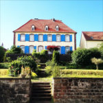 Kunterbuntes großes Haus frontal gesehen mit hellblauen Fensterläden und rotem Walmdach. Eine Treppe führt zwischen Sandsteinmauern auf das Gelände.