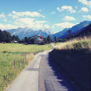 Blaustichig übersättigte Straßenszene einer offenbar bayerischen, ländlichen Dorfidylle mit Weg, der auf blaue Berge zuführt.