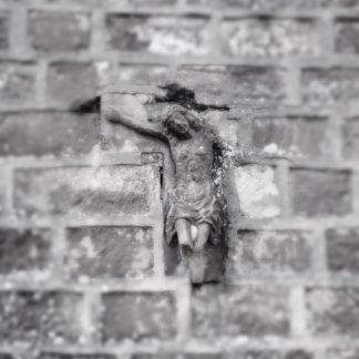 In eine Bruchsteinmauer ist ein halbes Jesuskreuz als Baustein integriert. Nur Kopf, Beine bis zum Knie und der rechte Arm sind noch erhalten.