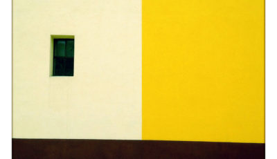 Dreiteilig rechtwinklig gefärbte Fassade. Links oben weiß mit Fenster, rechts oben gelb, unten zwei Streifen scharz und hellblau.
