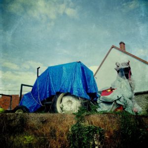Mit blauer Plane verdeckt steht ein Traktor vor dörflichem Wohnhaus. Das Bild ist Underfoot aufgenommen an einer dürr bewachsenen Böschung