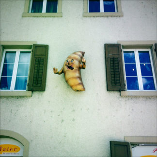 An einer Hauswand zwischen zwei Fenstern mit grünen Läden hängt ein personifiziertes, lachendes Croissant mit kleinen Ärmchen gestikulierend.