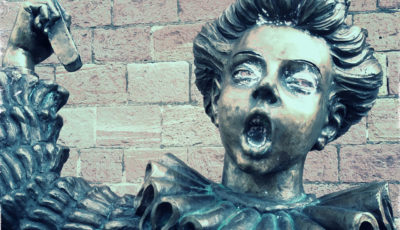 Oberkörper und Kopf einer Skulptur mit wallendem Gewand und gewelltem, großem Kragen vor eine blass-lila-braunen Sandsteinwand. Der Jüngling scheint laut zu singen, hat den Mund zum O geformt und reckt seinen rechten Arm.