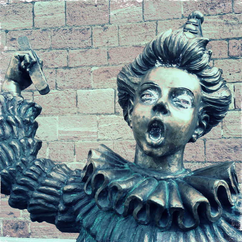 Oberkörper und Kopf einer Skulptur mit wallendem Gewand und gewelltem, großem Kragen vor eine blass-lila-braunen Sandsteinwand. Der Jüngling scheint laut zu singen, hat den Mund zum O geformt und reckt seinen rechten Arm.