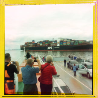 Eine handvoll Menschen mit dem Rücken zum Betrachter auf einer offenbaren Fähre, an der ein Containerschiff vorbeifährt. Die Menschen fotografieren das Containerschiff. Das quadratische Bild hat einen gelben Rand.