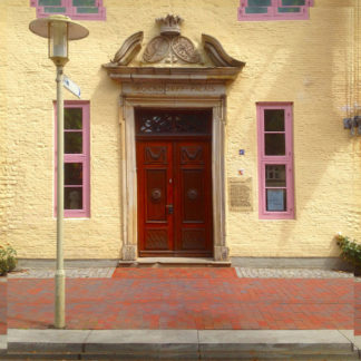 Eine verzeirte braune Tür in einer schmucken Hausfassade vor Gehweg neben Straßenlaterne. Zwei längliche hohe Fenster flankieren die Tür rechts und links. Der Gehweg besteht aus rotem Backsteinpflaster.