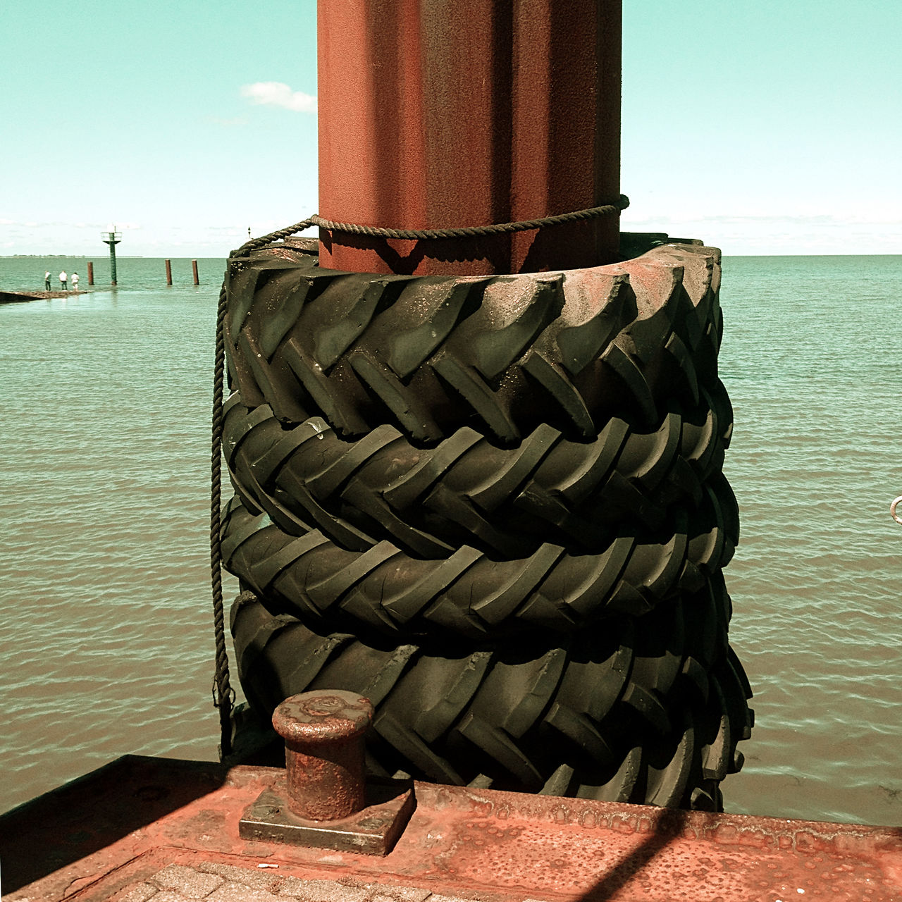 Vier alte Traktorhinterreifen umgeben einen rostogen Stahlpoller auf einem Schiffslandesteg. Dahinter das Meer und links ganz klein im Hintergrund weitere Landungsstege. Das Pfeilprofil der vier Reifen zeigt einheitlich nach links.