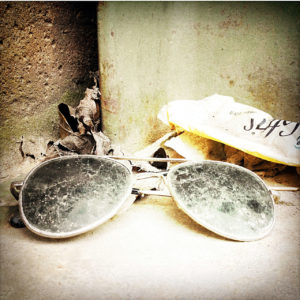 Eine schmutzige, zusammengeklappte Sonnebrille liegt in einer Ecke auf einer Betonmauer. Dahinter Papierfetzen und getrocknetes Laub.