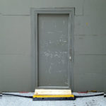 Sattgraue Tür in sattgrauer Messewand, davor ein querendes Kabel und eine gelbe Stufe