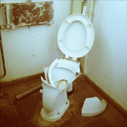 Eine geborstene, weiße Toilettenschüssel von davorstehend betrachtet. Der Deckel ist hochgeklappt. auf dem Boden liegt ein Porzellanstück. Das WC ist in einer Ecke vor alten dicken Rohren mit klassischer Druckspülung montiert.