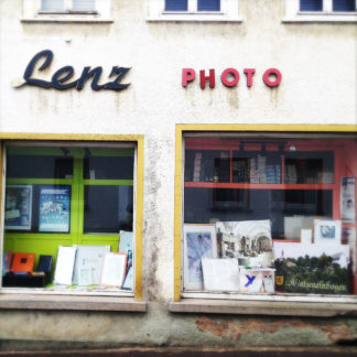Fassade eines innerstädtsischen Fotoladens mit schöner alter Schrift Lenz PHOTO. Das H im Schriftzug wurde notdürftig repariert. Zwei Schaufenster mit gerahmten Bildern.