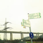 Weiße Hinweisschilder mit grüner Schrift an einem Pfosten. Radwege. Im Hintergrund eine Schnellstraßenbrücke und ein Strommast mit Hochspannungsleitung.