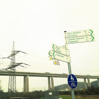 Weiße Hinweisschilder mit grüner Schrift an einem Pfosten. Radwege. Im Hintergrund eine Schnellstraßenbrücke und ein Strommast mit Hochspannungsleitung.