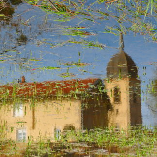 Spiegelung einer burgundischen Dorfkirche in einem mit Gras und Schilf durchsetzten Tümpel. Das Bild steht kopf und man sieht die Kirche richtig herum vor blauem Himme unter gekräuselten Wellen. Ein einfaches Kirchenschiff mit rotem Dach und kleinem Kuppelturm.
