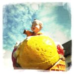 Underfootaufnahme eines übergroßen gelben Eisbällchens, auf dem eine Kinderfigur die abe Hand gen Himmel reckt.