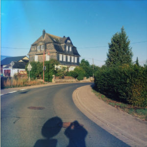 Straßenflucht auf ein großes einzelstehendes Haus mit Walmdach unter blauem Himmel. Am Rand Hecken und ein Baum. Der Schatten des Fotografen fällt neben dem Schatten eines runden Verkehrsschilds auf die Straße.
